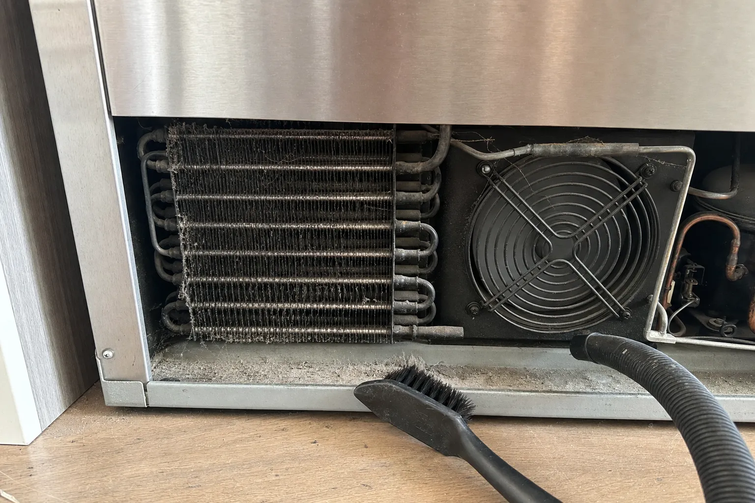 Dust-clogged condenser coil and fan behind the open base grille of a stainless refrigerator, brush and vacuum hose alongside